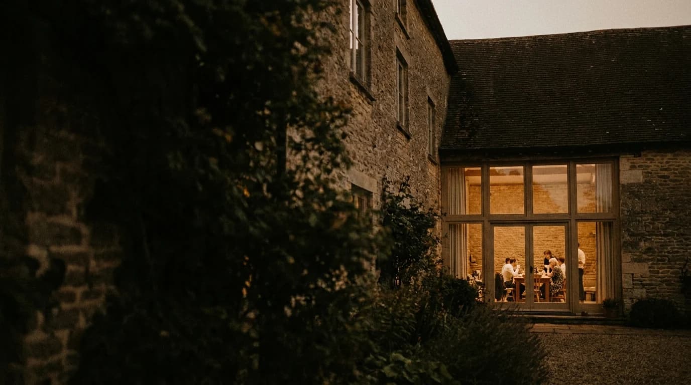 A stone house at dusk, seen through the garden, with a small group gathered at a table in a lit room